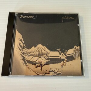 May include: A Weezer "Pinkerton" CD in a clear jewel case. The album art features a snowy landscape with mountains, buildings, and figures. The band's name, "Weezer", is written in white, and the album title, "Pinkerton", is in a script font.