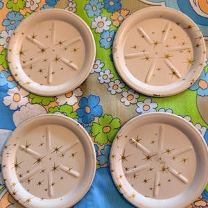 May include: Four white metal coasters with a gold starburst design. Each coaster has a raised rim and six radial support lines. The coasters are arranged on a floral patterned fabric with blue, green, and white flowers.