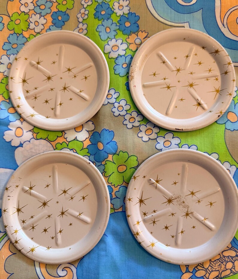 May include: Four white metal coasters with a gold starburst design. Each coaster has a raised rim and six radial support lines. The coasters are arranged on a floral patterned fabric with blue, green, and white flowers.