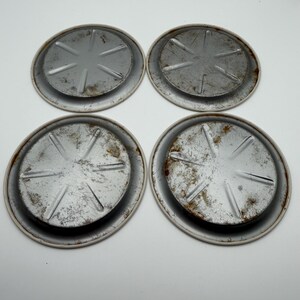 May include: Four round, metal coasters with a distressed, aged appearance. Each coaster has a raised center with a starburst design and a white rim. The metal shows signs of rust and wear, giving them a vintage aesthetic.