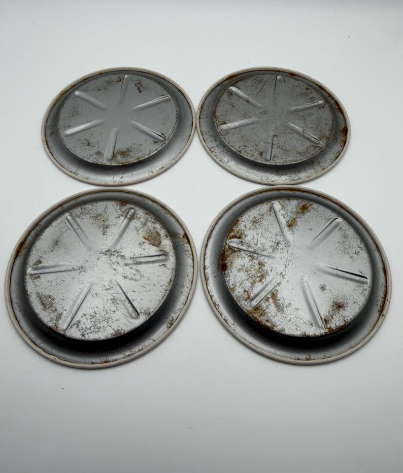May include: Four round, metal coasters with a distressed, aged appearance. Each coaster has a raised center with a starburst design and a white rim. The metal shows signs of rust and wear, giving them a vintage aesthetic.