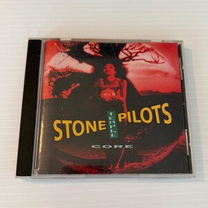 May include: A compact disc case featuring the album "Core" by Stone Temple Pilots. The cover art displays a red and black background with a figure holding an orb. The band's name is in yellow, with "Temple" in green.