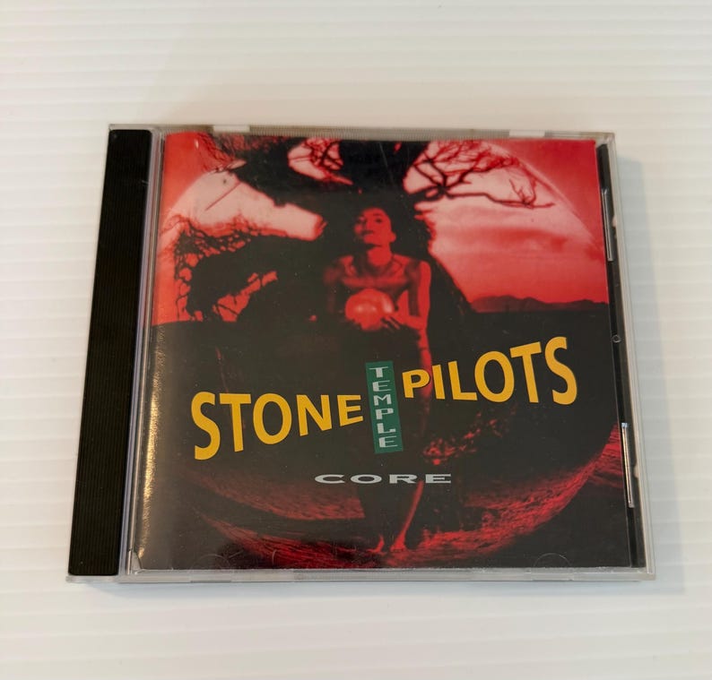 May include: A compact disc case featuring the album "Core" by Stone Temple Pilots. The cover art displays a red and black background with a figure holding an orb. The band's name is in yellow, with "Temple" in green.