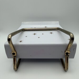 May include: A vintage white serving dish with a gold-colored handle and starburst pattern. The dish has a rectangular shape and is supported by a gold-colored metal frame.