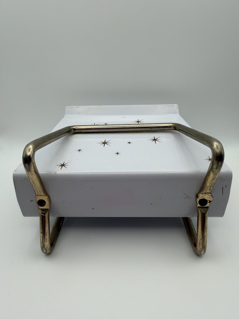 May include: A vintage white serving dish with a gold-colored handle and starburst pattern. The dish has a rectangular shape and is supported by a gold-colored metal frame.