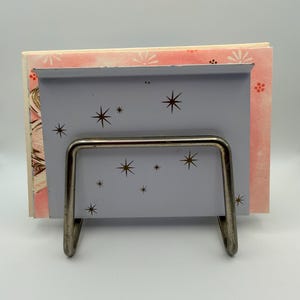May include: A vintage gray metal book holder with a silver wire frame. The holder features a starburst design and holds a stack of books with pink and white floral patterned covers. The overall aesthetic is retro and mid-century modern.