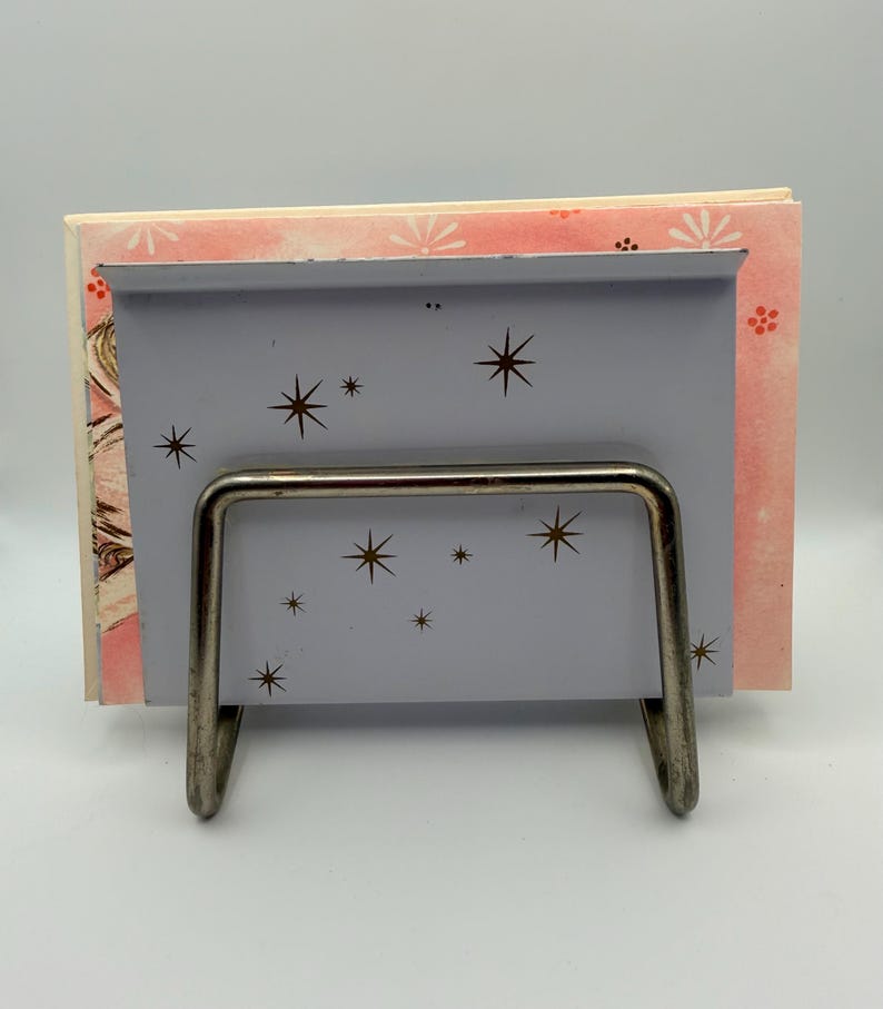 May include: A vintage gray metal book holder with a silver wire frame. The holder features a starburst design and holds a stack of books with pink and white floral patterned covers. The overall aesthetic is retro and mid-century modern.