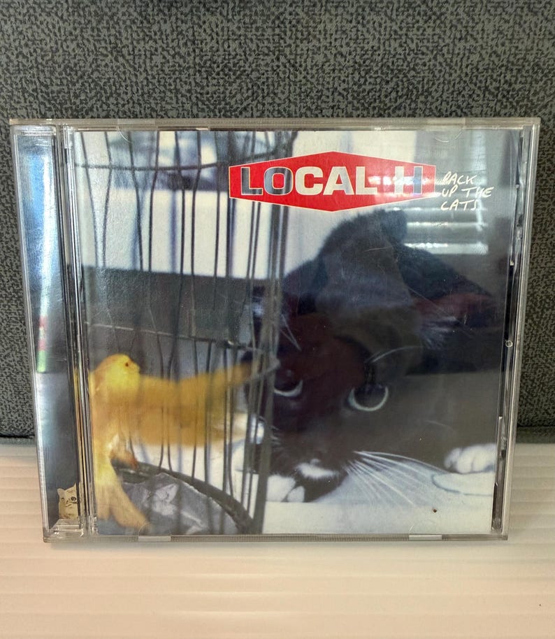 May include: A CD case featuring the album "Back Up the Cats" by Local H. The cover art shows a yellow bird in a cage and a black and white cat. The band's name is in a red and white logo.