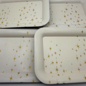 May include: Four rectangular serving trays with rounded corners. The trays are off-white with a gold starburst pattern. The trays are stacked together, with the top tray showing some wear.
