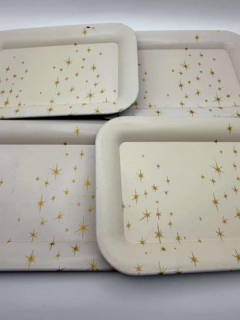 May include: Four rectangular serving trays with rounded corners. The trays are off-white with a gold starburst pattern. The trays are stacked together, with the top tray showing some wear.