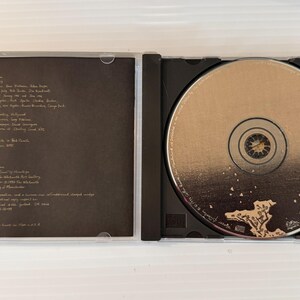May include: Open CD case with a disc inside. The disc is gold with a central hub and text around the edges. The left side of the case has a dark brown insert with white text. The case is clear plastic.