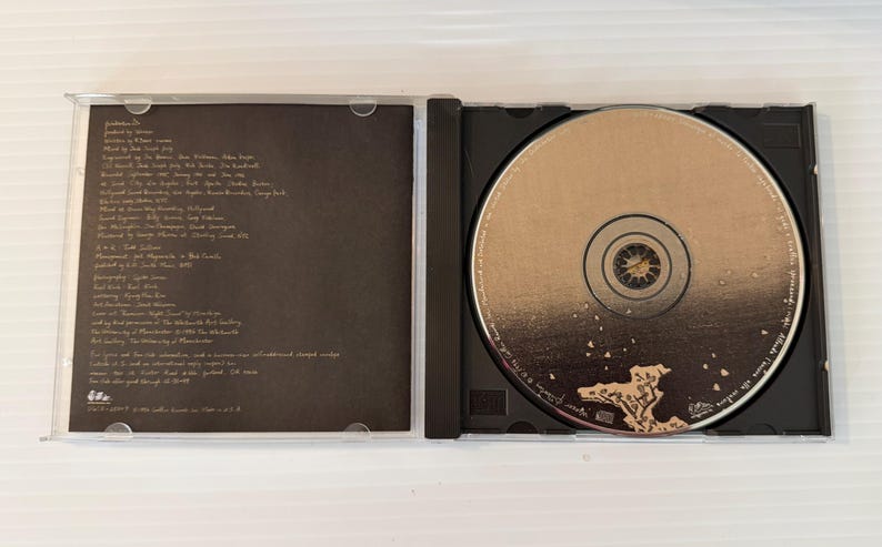 May include: Open CD case with a disc inside. The disc is gold with a central hub and text around the edges. The left side of the case has a dark brown insert with white text. The case is clear plastic.