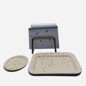 May include: A set of vintage serving trays and a notepad holder. The trays are off-white with a gold starburst pattern. The notepad holder has a matching design. The metal stand is a warm, brassy color.