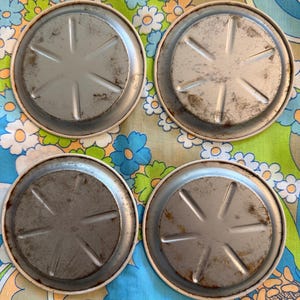 May include: Four vintage metal coasters with a silver-colored center and a white rim. The coasters have a starburst design on the bottom and show signs of wear. The coasters are arranged on a colorful floral patterned fabric.