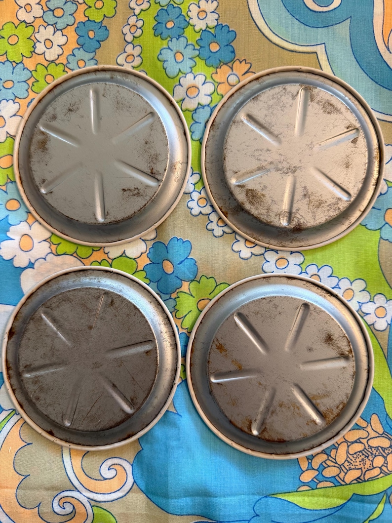 May include: Four vintage metal coasters with a silver-colored center and a white rim. The coasters have a starburst design on the bottom and show signs of wear. The coasters are arranged on a colorful floral patterned fabric.