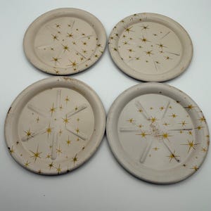 May include: Four off-white coasters with a gold starburst design. Each coaster has a slightly raised edge and a series of gold stars radiating from the center. The coasters are arranged in a square formation.