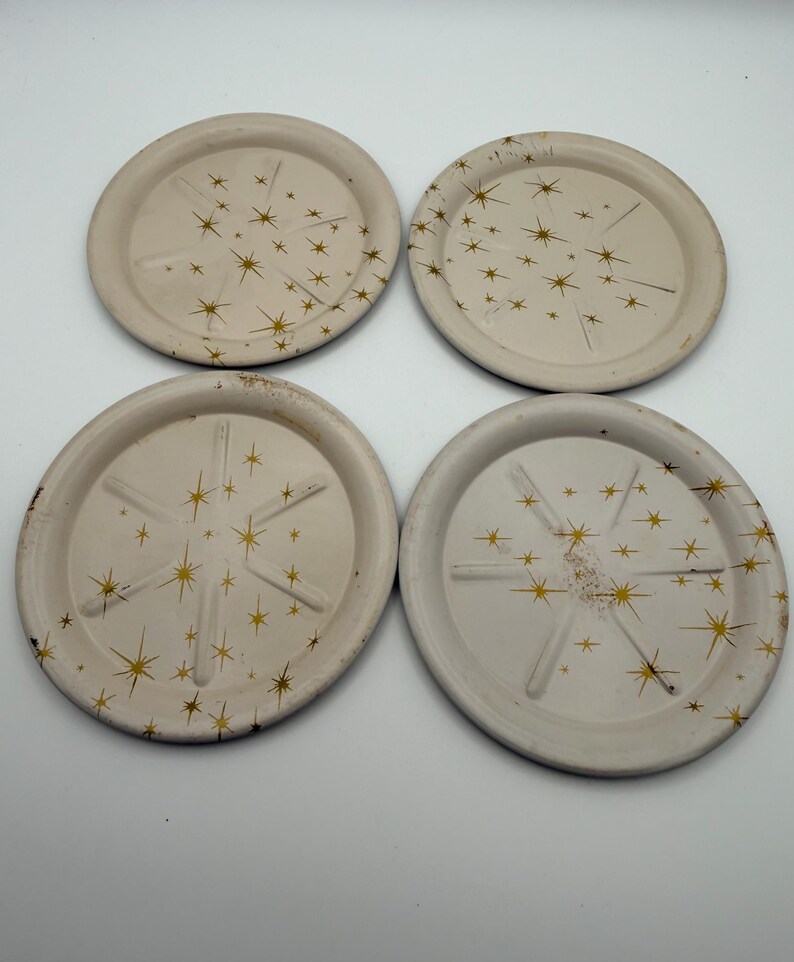 May include: Four off-white coasters with a gold starburst design. Each coaster has a slightly raised edge and a series of gold stars radiating from the center. The coasters are arranged in a square formation.