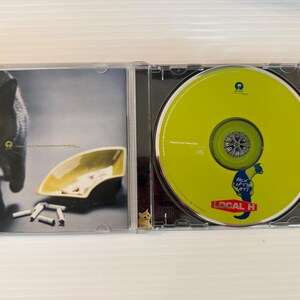 May include: A CD case is open, revealing a lime green CD with the text "LOCAL H" and a cartoon seahorse. The left side shows a black cat, an ashtray, and cigarettes.