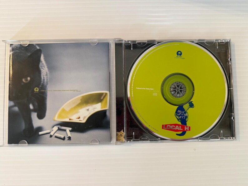 May include: A CD case is open, revealing a lime green CD with the text "LOCAL H" and a cartoon seahorse. The left side shows a black cat, an ashtray, and cigarettes.