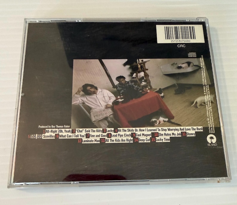 May include: A compact disc case with a black background and a photograph of a room with people and a table. The back of the CD case has a tracklist with song titles and the text "ISLAND".