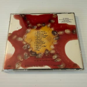 May include: A CD case featuring a colorful design with a red and yellow background and a pattern of seeds. The tracklist includes titles like "Soak the Sun" and "Seed to a Tree". The CD case is in a clear plastic case.