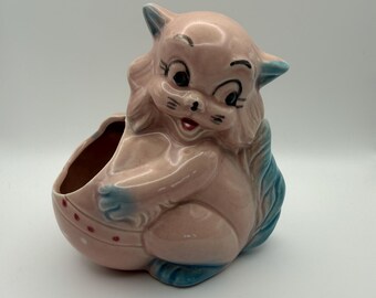 May include: A vintage ceramic planter in the shape of a cartoon cat. The planter is primarily pink with blue accents on the ears, paws, and tail. The cat has black eyes, whiskers, and a red mouth. The planter has a small opening for plants.