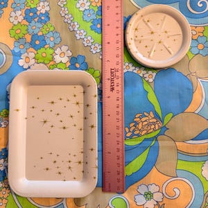 May include: A white rectangular tray and a small round coaster with gold starburst designs. A pink ruler shows the tray is approximately 8 inches long. The objects are on a colorful floral patterned fabric.