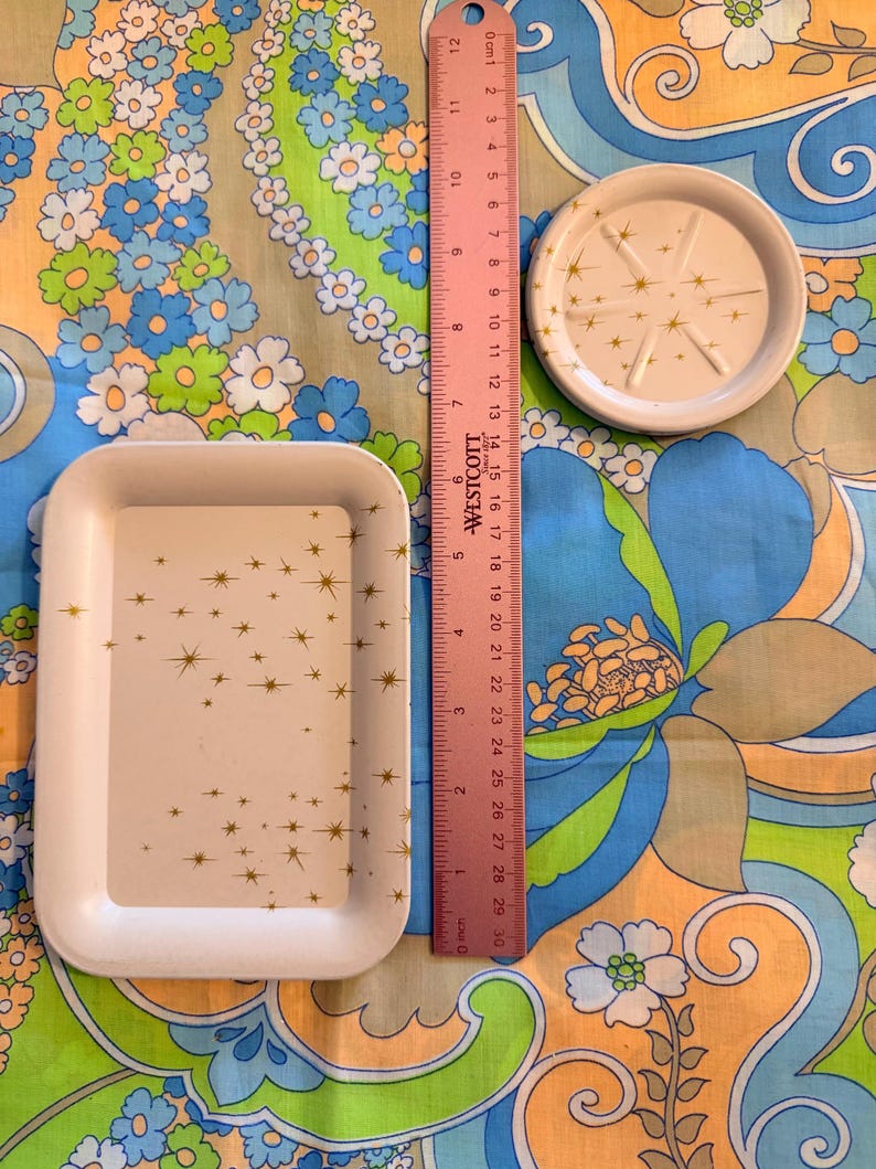 May include: A white rectangular tray and a small round coaster with gold starburst designs. A pink ruler shows the tray is approximately 8 inches long. The objects are on a colorful floral patterned fabric.