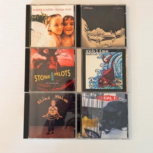 May include: A collection of six CD albums in jewel cases. The album covers feature various designs, including band names and album titles. The albums include Smashing Pumpkins, Weezer, Stone Temple Pilots, Sublime, Blind Melon, and Local H.