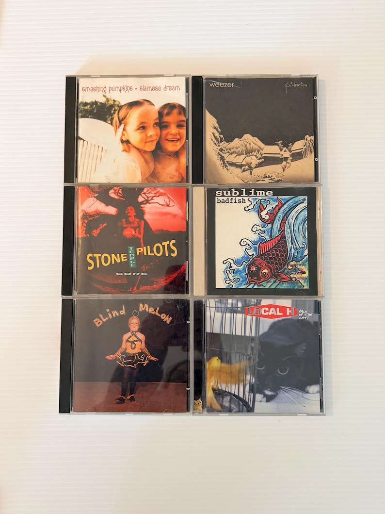 May include: A collection of six CD albums in jewel cases. The album covers feature various designs, including band names and album titles. The albums include Smashing Pumpkins, Weezer, Stone Temple Pilots, Sublime, Blind Melon, and Local H.