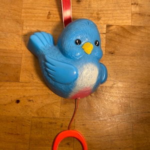 May include: A blue plastic bird-shaped toy with a yellow beak, black eyes, and a red pull ring. The bird has white and pink accents on its chest and belly. A red ribbon is attached to the top.