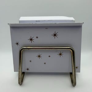 May include: A white metal card holder with a mid-century modern design. The holder features a series of brown starbursts and a silver metal stand. The holder is designed to hold cards or notes.