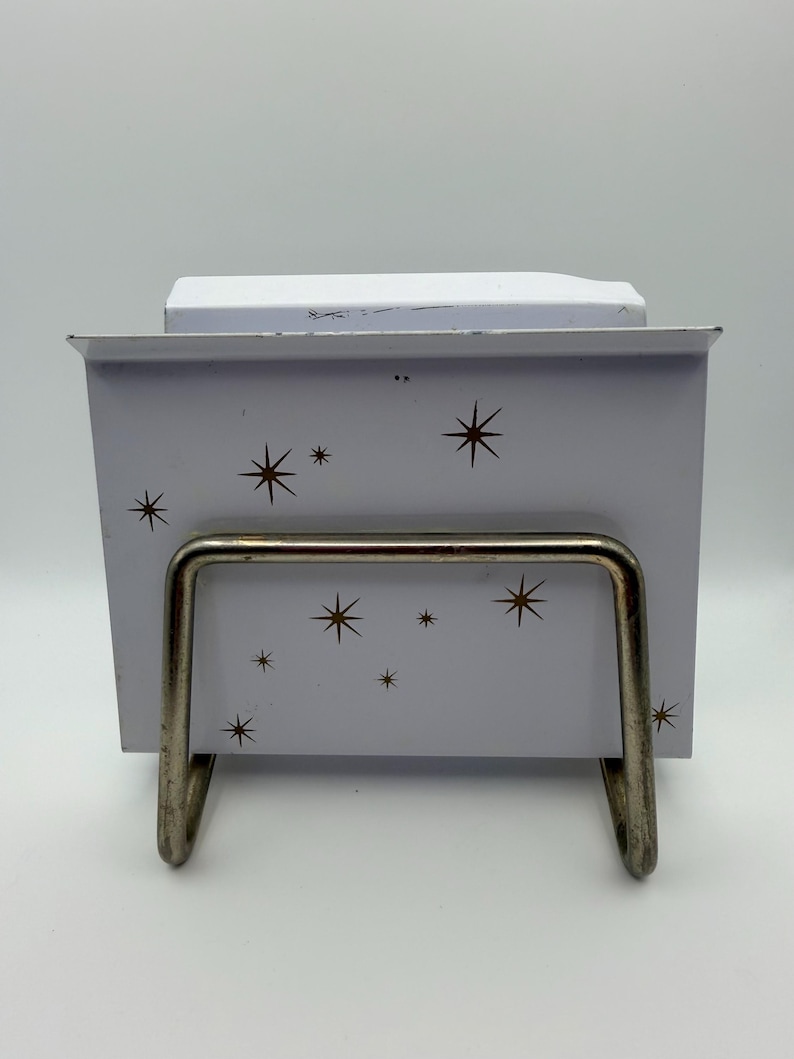 May include: A white metal card holder with a mid-century modern design. The holder features a series of brown starbursts and a silver metal stand. The holder is designed to hold cards or notes.