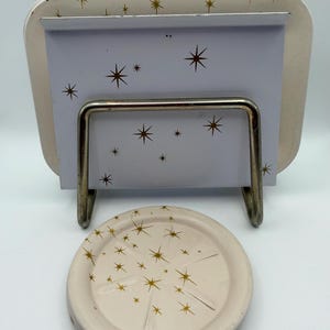 May include: A cream-colored metal tray and coaster set with a mid-century modern design. The tray has a rectangular shape with rounded corners and a metal stand. Both pieces feature a gold starburst pattern.