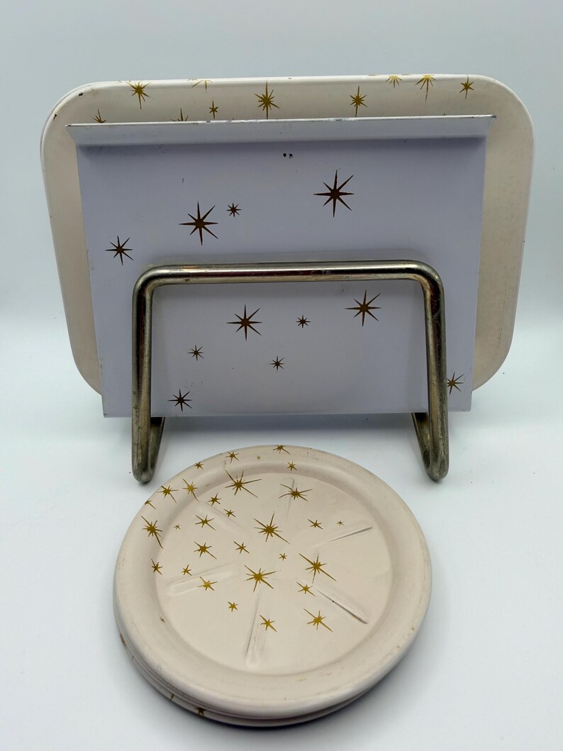 May include: A cream-colored metal tray and coaster set with a mid-century modern design. The tray has a rectangular shape with rounded corners and a metal stand. Both pieces feature a gold starburst pattern.