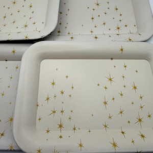 May include: A collection of off-white rectangular trays with rounded corners. Each tray is decorated with a gold starburst pattern. The trays are stacked, showcasing their design and potential for serving or display.