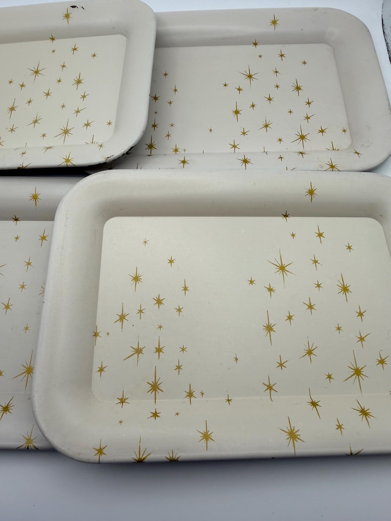 May include: A collection of off-white rectangular trays with rounded corners. Each tray is decorated with a gold starburst pattern. The trays are stacked, showcasing their design and potential for serving or display.