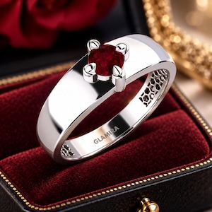 Peut inclure: Une bague en métal argenté poli ornée d'une pierre précieuse rouge foncé de taille ronde fixée par un sertissage à quatre griffes. L'anneau présente une surface large et lisse avec le nom de marque GLAMIRA gravé à l'intérieur de la bande.