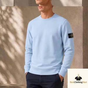 May include: A light blue long-sleeve sweatshirt with a crew neckline and ribbed cuffs. The garment features a black and yellow circular brand patch on the left sleeve. The sweatshirt is paired with dark navy blue trousers.