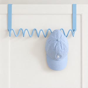 May include: A light blue over-the-door metal rack with a wavy, serpentine design featuring multiple hooks for storage. A matching light blue baseball cap with a small white embroidered logo is hanging on one of the hooks of the rack.