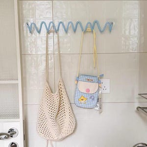 May include: A light blue spiral wall hook rack holding a cream-colored crocheted net tote bag and a small light blue and white gingham crossbody bag with a yellow star patch, a pink circular button, and a thin yellow shoulder strap.