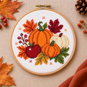 Op de afbeelding: Een ronde houten borduurring met een kruissteekpatroon van twee oranje pompoenen, een witte pompoen, een rode appel en diverse herfstbladeren in tinten oranje, rood en groen, geaccentueerd met kleine rode bessen op een witte ondergrond.