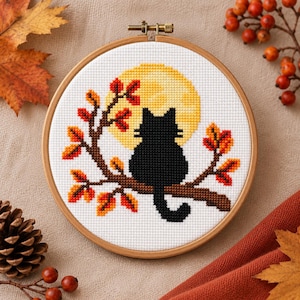 Puede incluir: Un bastidor de bordado circular de madera que contiene un diseño de punto de cruz de un gato negro sentado en una rama de árbol marrón. El gato se recorta contra una gran luna llena amarilla, rodeado de hojas de otoño naranjas y rojas.