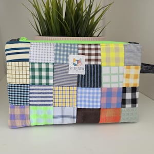 Puede incluir: Un estuche rectangular con cremallera hecho de un patchwork de varios cuadrados de tela con diversos patrones, incluyendo cuadros vichy, rayas y colores sólidos. Una etiqueta blanca con un icono de gato y el texto PITAPITURRA está centrada.