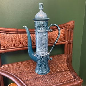 May include: A tall, cylindrical ceramic teapot in shades of teal and blue, featuring a textured, woven-like pattern on the base and neck, a smooth spout, a curved handle, and a matching lid with a small, rounded finial on top.