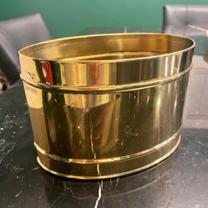 Peut inclure: Un cache-pot ovale en laiton avec une finition métallique polie et réfléchissante. Le récipient présente une surface lisse et continue avec une bande horizontale en relief entourant le milieu, offrant un aspect doré brillant.