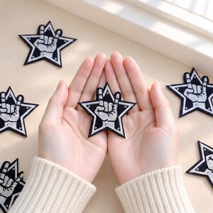 Puede incluir: Una colección de parches bordados en forma de estrella con fondo negro, borde blanco y un gráfico central de una mano blanca haciendo el signo de rock and roll. Los parches están dispuestos de forma dispersa sobre una superficie clara.