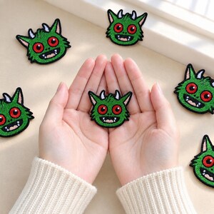 May include: A collection of embroidered patches featuring a green monster head with large red eyes, white horns, and a wide mouth with visible teeth. The patches are circular in shape with black borders and are arranged on a light surface.