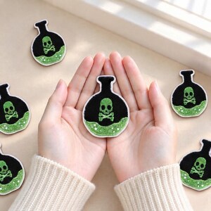 Könnte beinhalten: Mehrere gestickte Aufnäher in Form von runden Zaubertrankflaschen mit schwarzem Stoff, einem grünen Totenkopf-mit-gekreuzten-Knochen-Motiv und einer grünen Flüssigkeitsbasis. Die Aufnäher haben einen weißen Rand und liegen auf einer hellen Fläche.