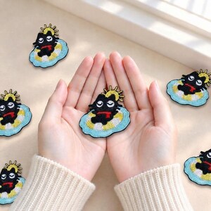 May include: Embroidered patches featuring a black cat with a red scarf resting on a yellow and white striped inner tube, surrounded by a light blue water shape and a yellow sun icon, arranged in a repeating pattern across the frame.
